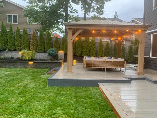 Wooden gazebo with string lights over patio and lawn in landscaped backyard.