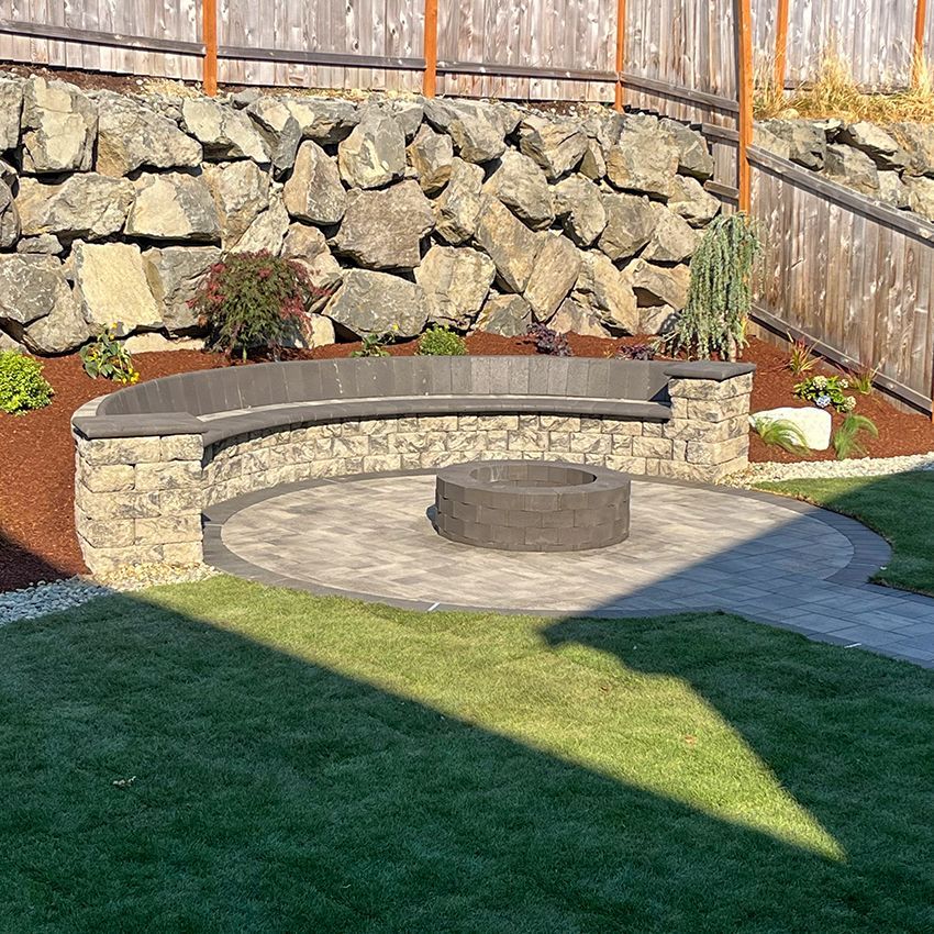Stone patio with fire pit, curved bench, rock wall backdrop, and grass lawn.