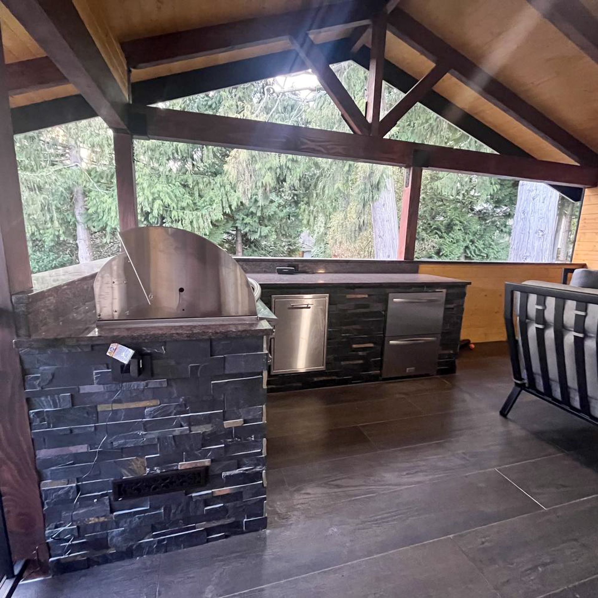 Covered outdoor kitchen with stone grill, black countertop, and wooded view under a wooden roof.