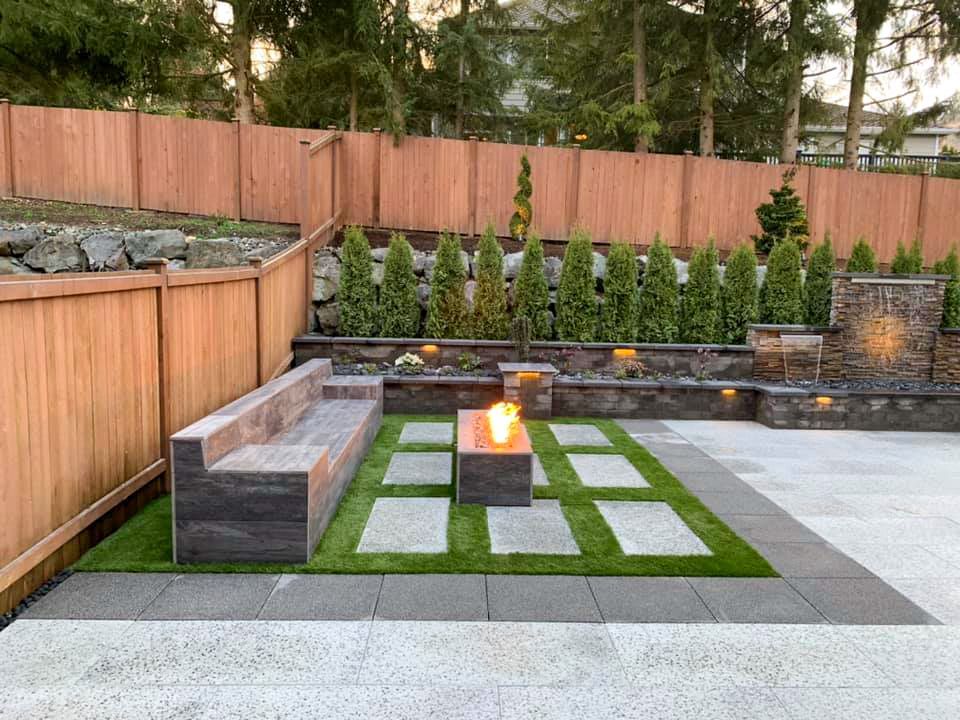 Modern patio with stone bench and fire pit surrounded by greenery and wooden fence.