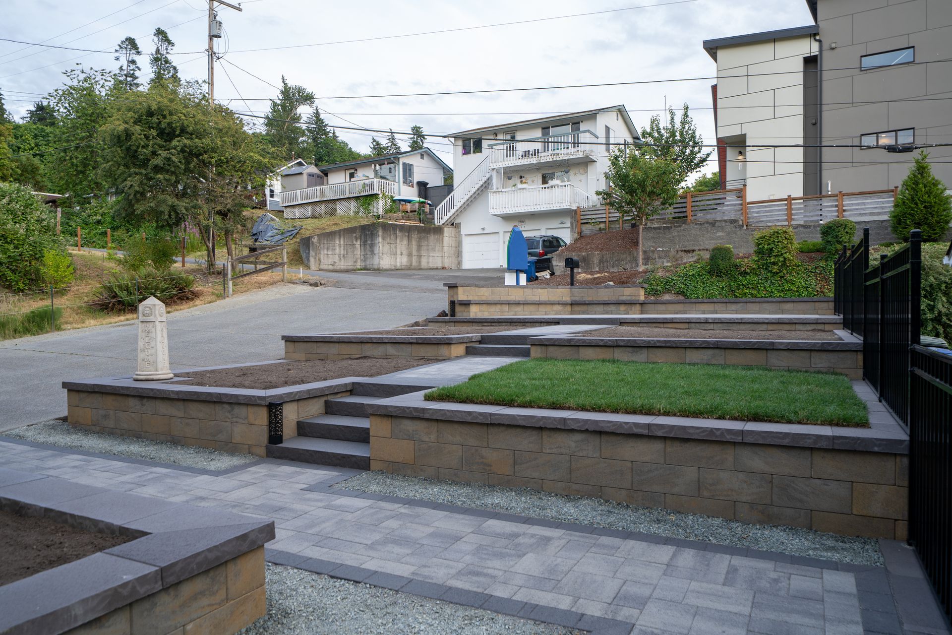 Stone tiered garden with grass and plants; paved area with buildings on a hill in the background.