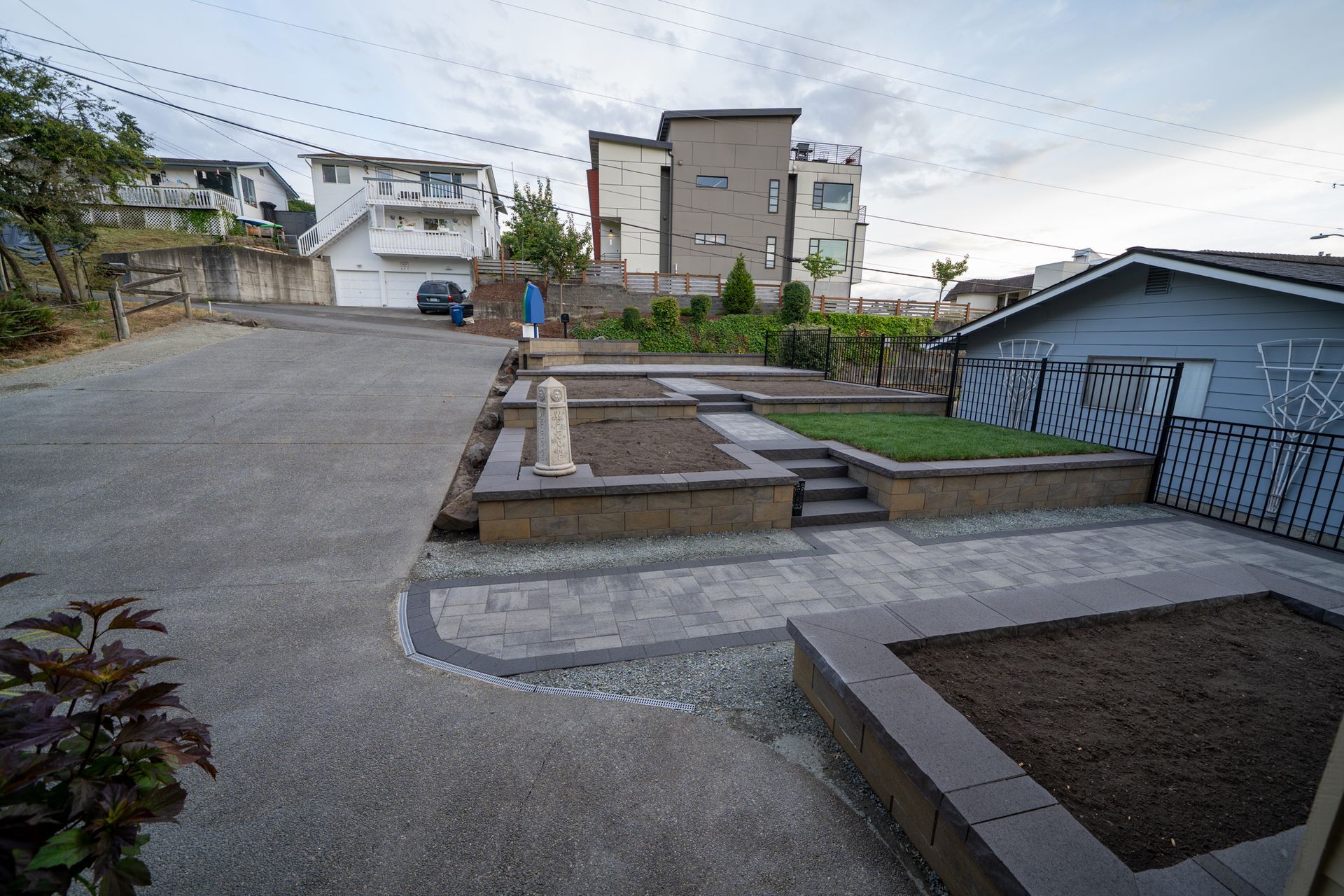 A concrete driveway leads to a tiered garden with raised beds and buildings in the background under a cloudy sky.