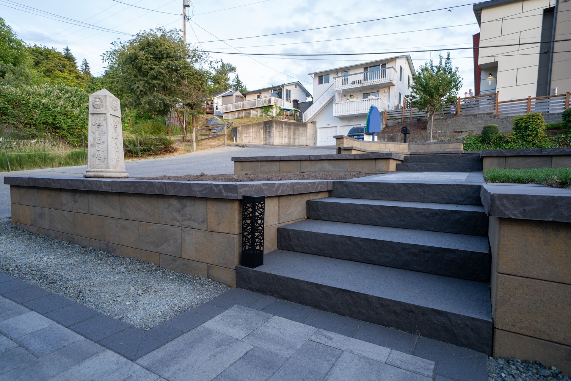 Stone steps and retaining wall leading up to a hillside with houses, featuring a decorative light post.