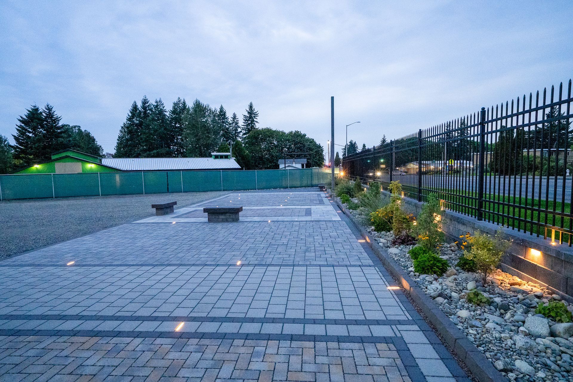 Brick paved parking area with landscaping, lit by small ground lights, bordered by a black fence.