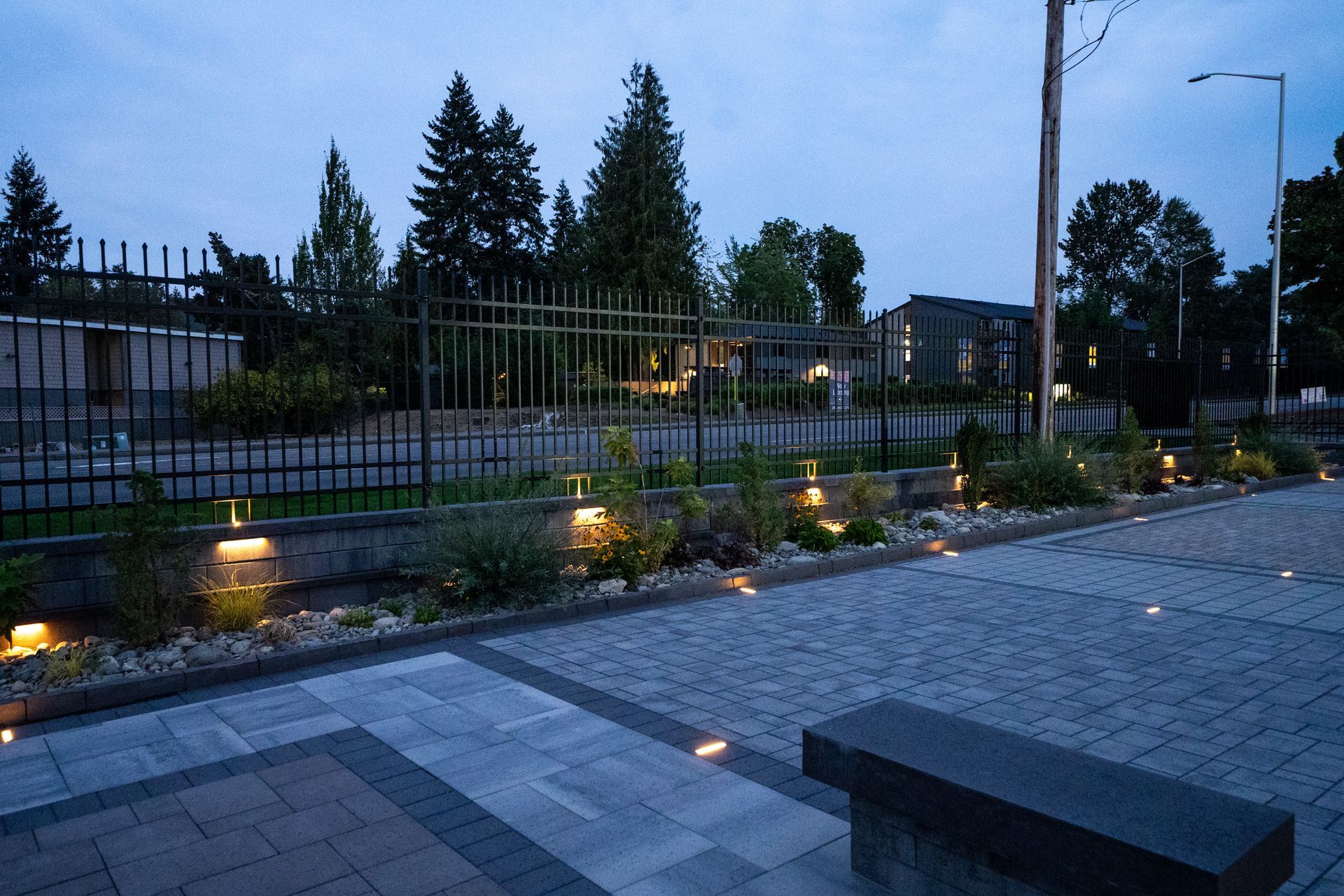 Nighttime exterior scene with lit garden beds, paving stones, and a bench.