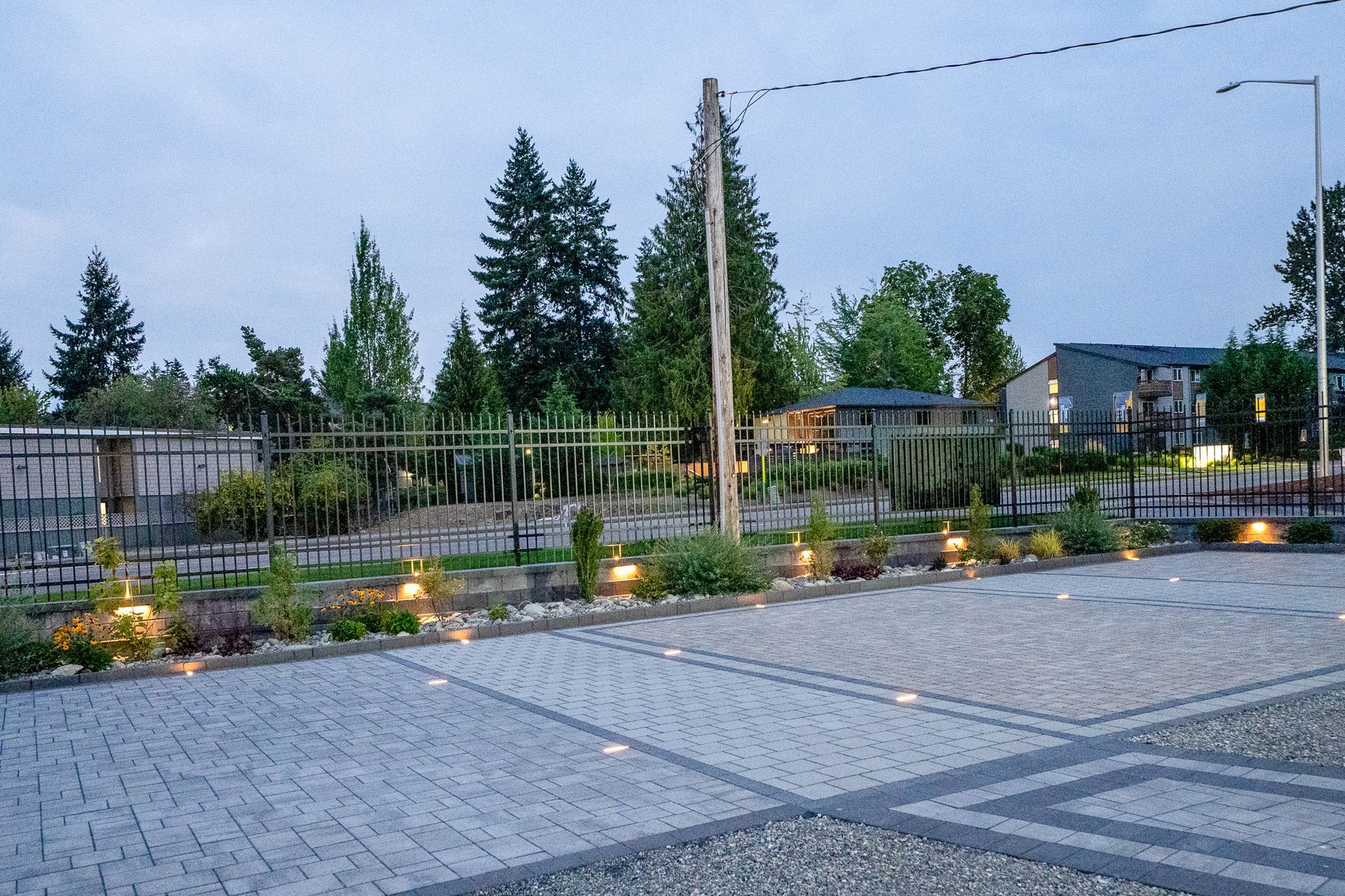Evening view of a paved area with in-ground lights, fence, landscaping, and buildings in the background.
