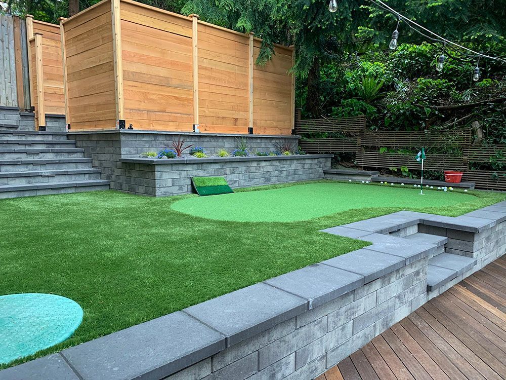 A backyard putting green with stone retaining walls, stairs, and a wooden privacy fence.