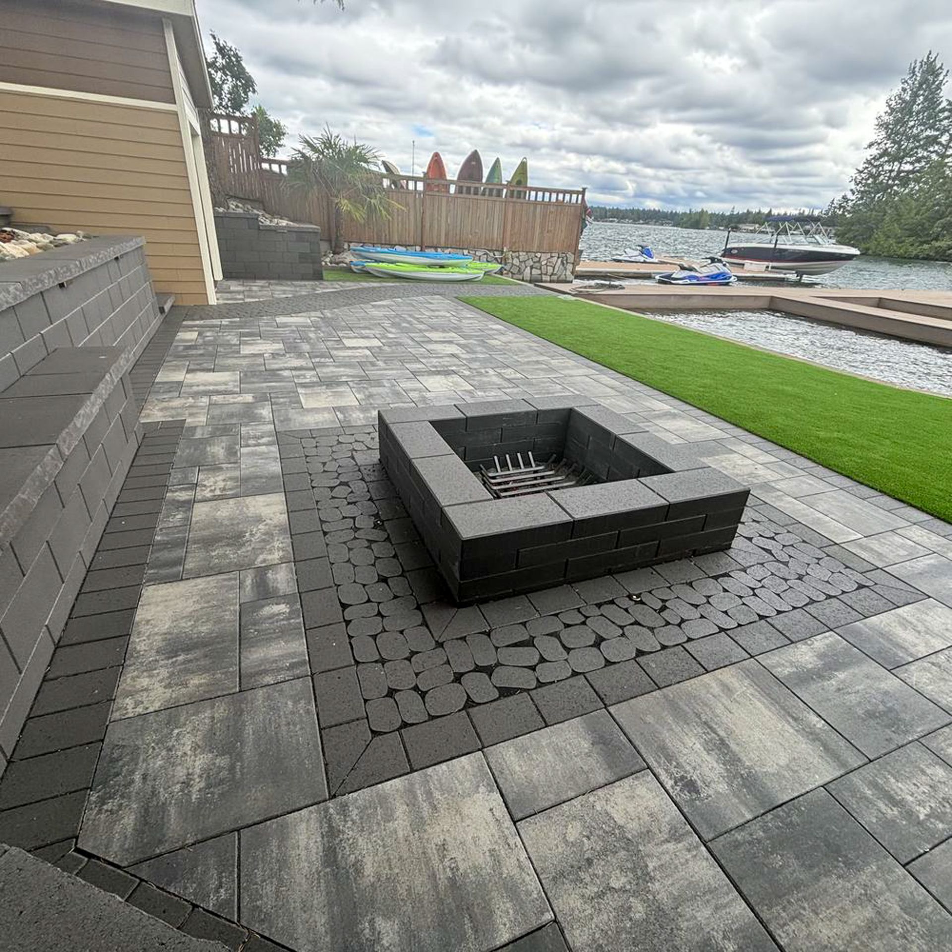 Patio with fire pit near a lake. Pavers in gray and black, green grass, kayaks, cloudy sky.
