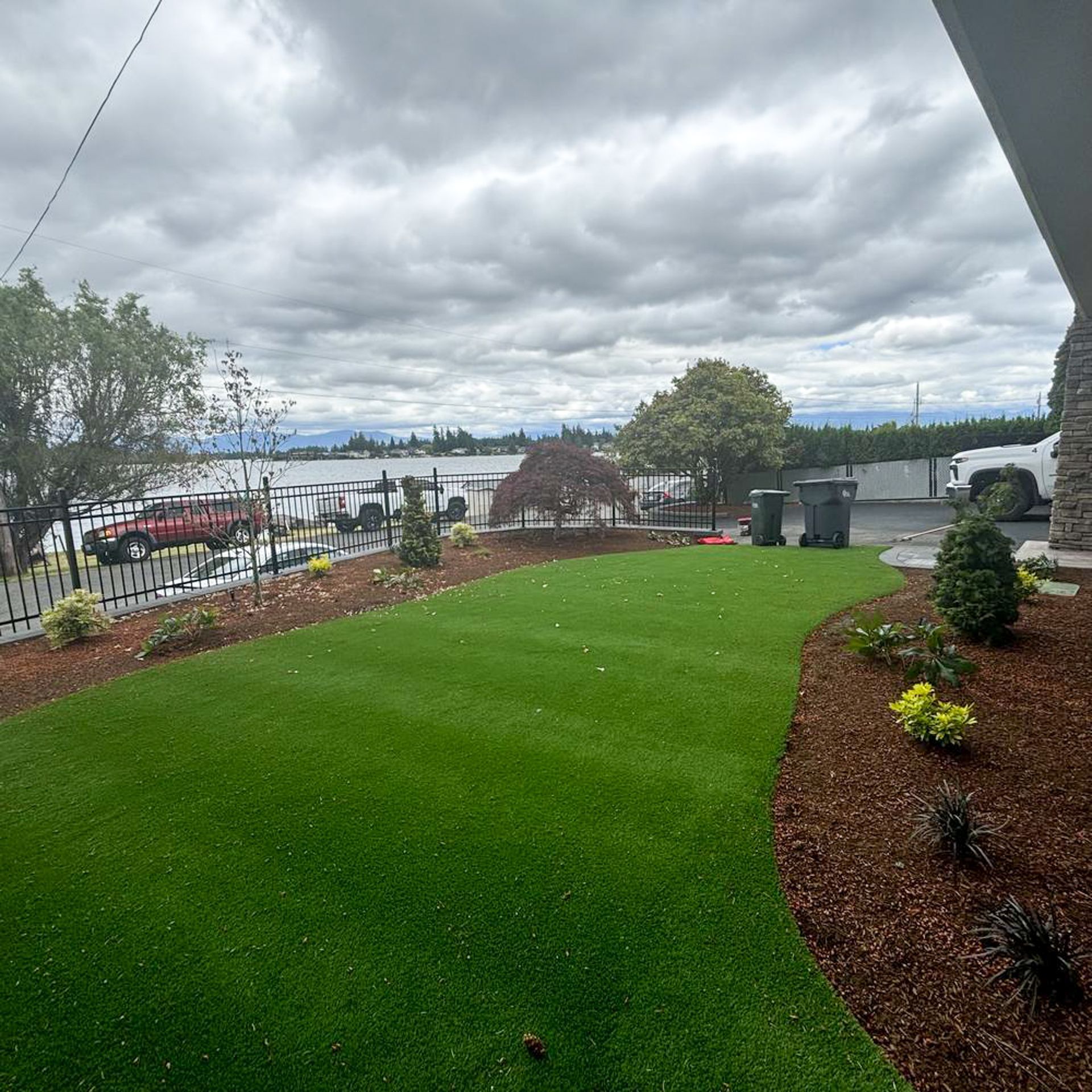 Lush green artificial turf lawn with a mulched flowerbed against a cloudy sky.