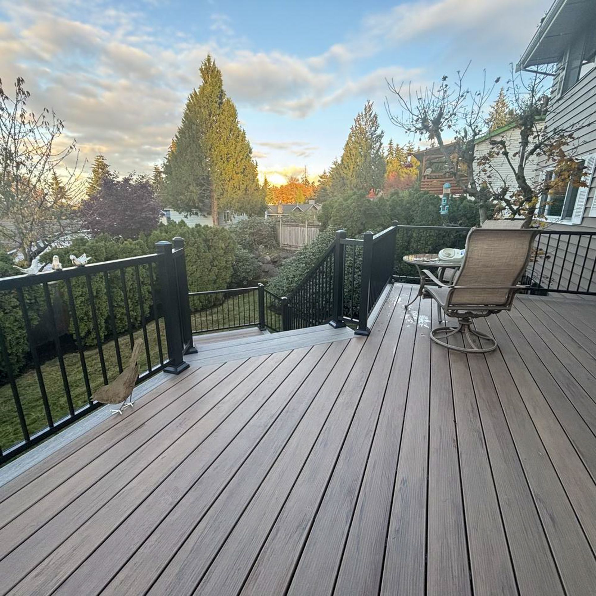 Wooden deck with black railing and a table/chair overlooking a backyard with trees.