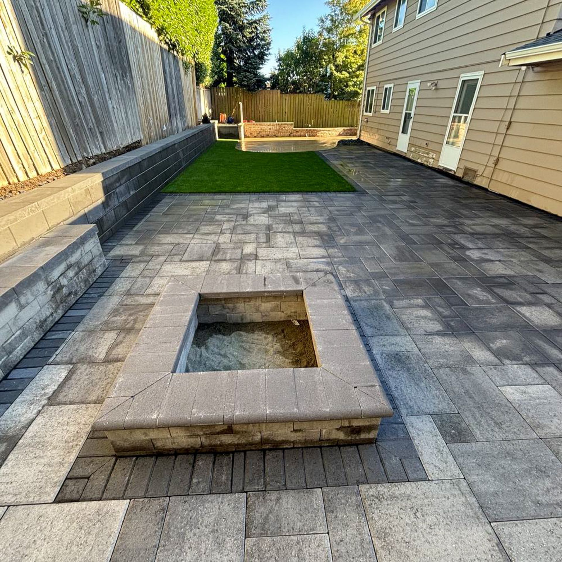Patio with fire pit, gray pavers, small lawn, and beige house.