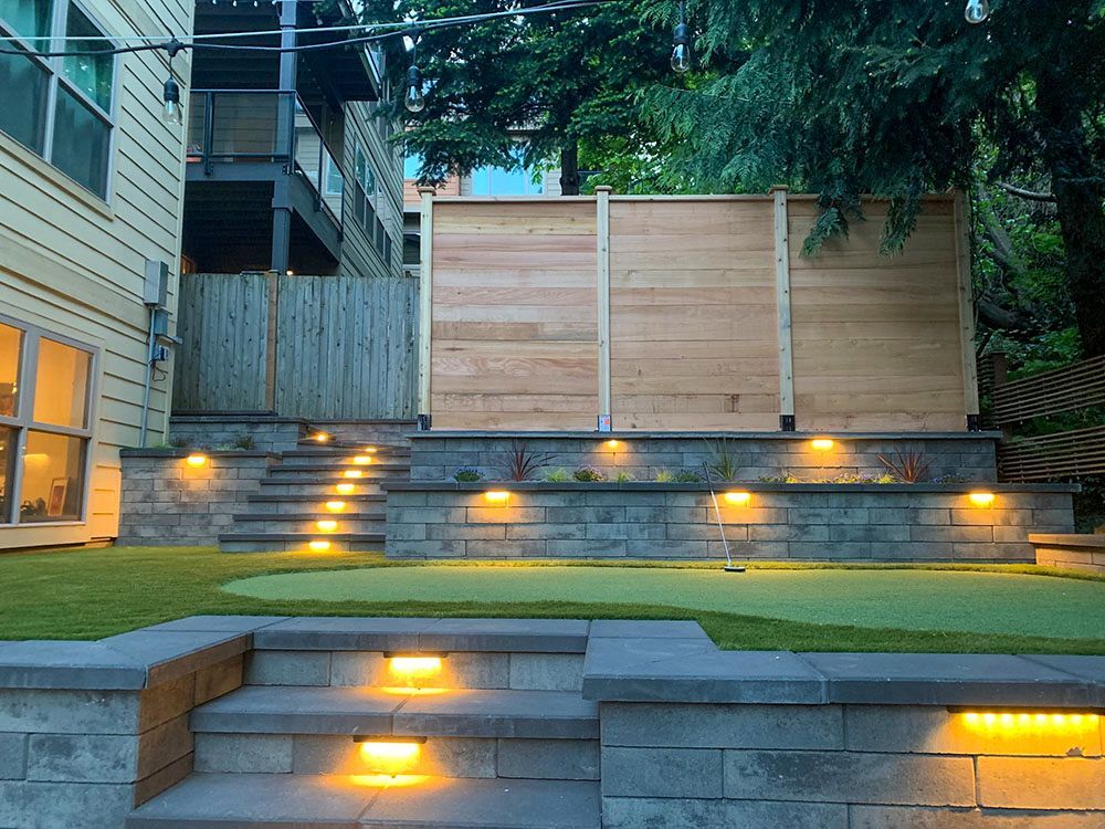 Backyard with lit steps, stone walls, a small putting green, and wood privacy fence.