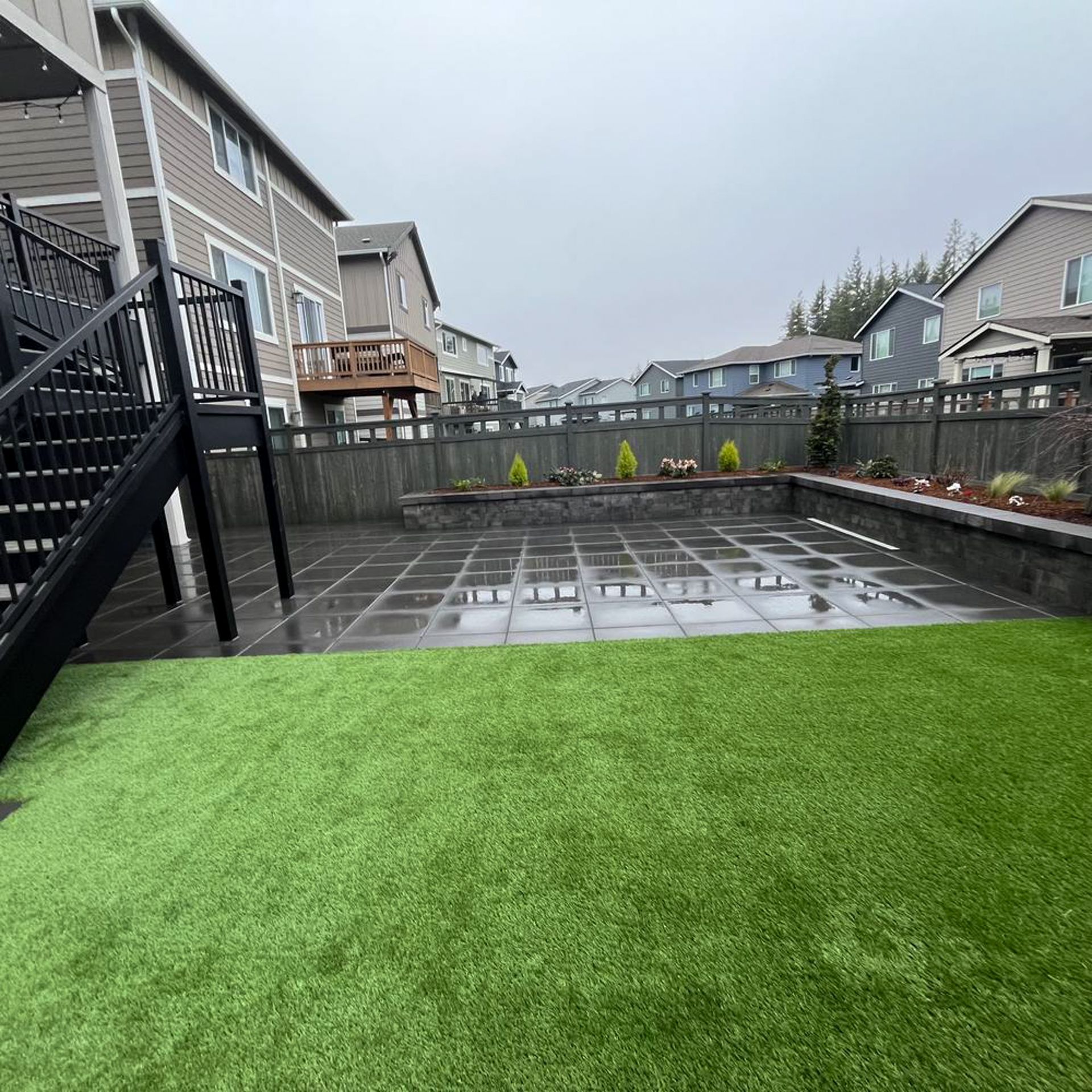 Backyard with artificial grass, patio, retaining wall, wooden fence, and multi-story houses.