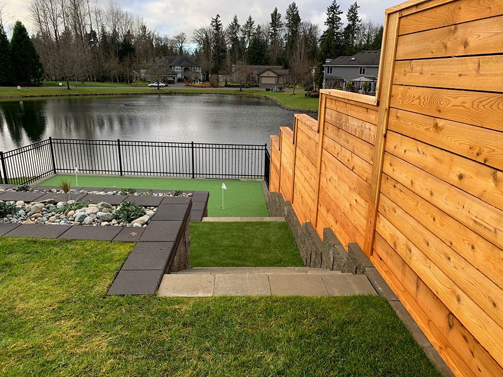 A backyard golf green next to a lake, with a wooden fence and stone steps.