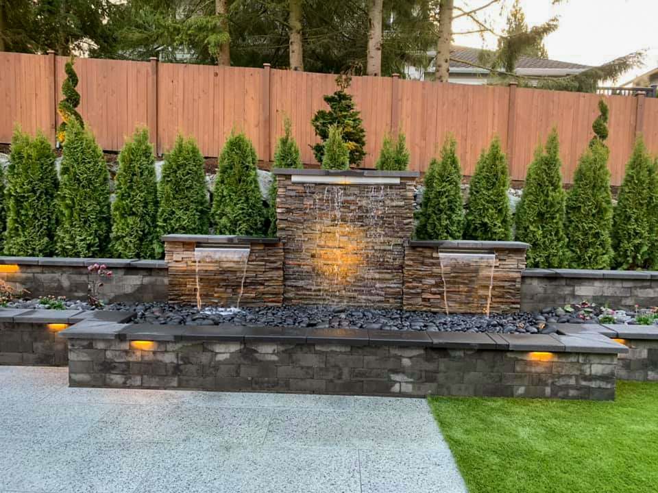 Water fountain with stone facade, surrounded by green shrubs, brick walls, and a wooden fence.