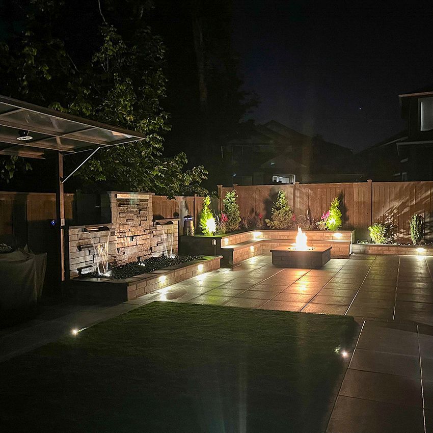 Nighttime outdoor patio with fire pit, water feature, built-in seating, and landscape lighting.