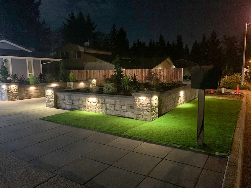Night view of a well-lit house with stone retaining walls, green grass, and landscaping with a dark sky background.