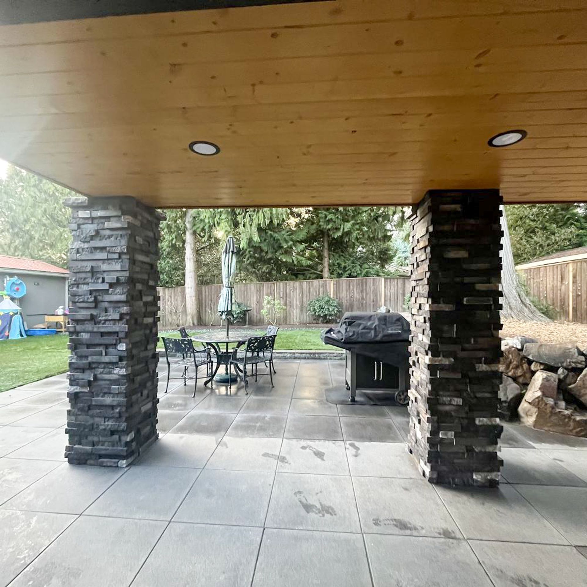 Patio with stone pillars, grill, table, and backyard view with trees.