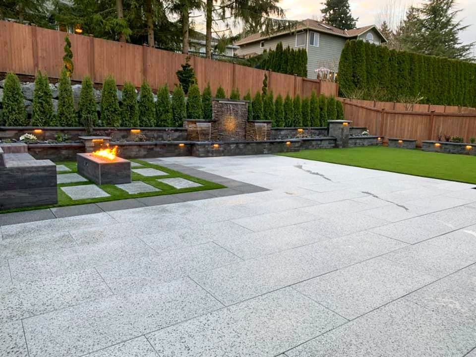 Backyard patio with fire pit, water feature, and wooden fence. Green grass and gray paving stones.