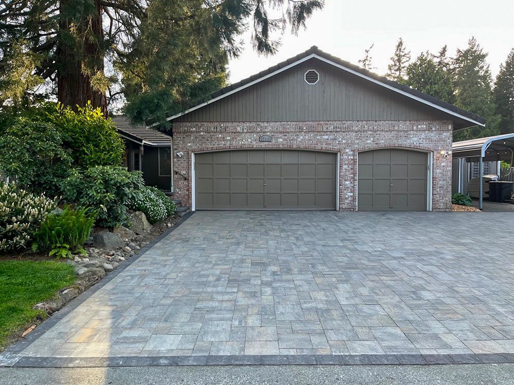 Brick garage with two doors, gray paver driveway, and greenery on the left.