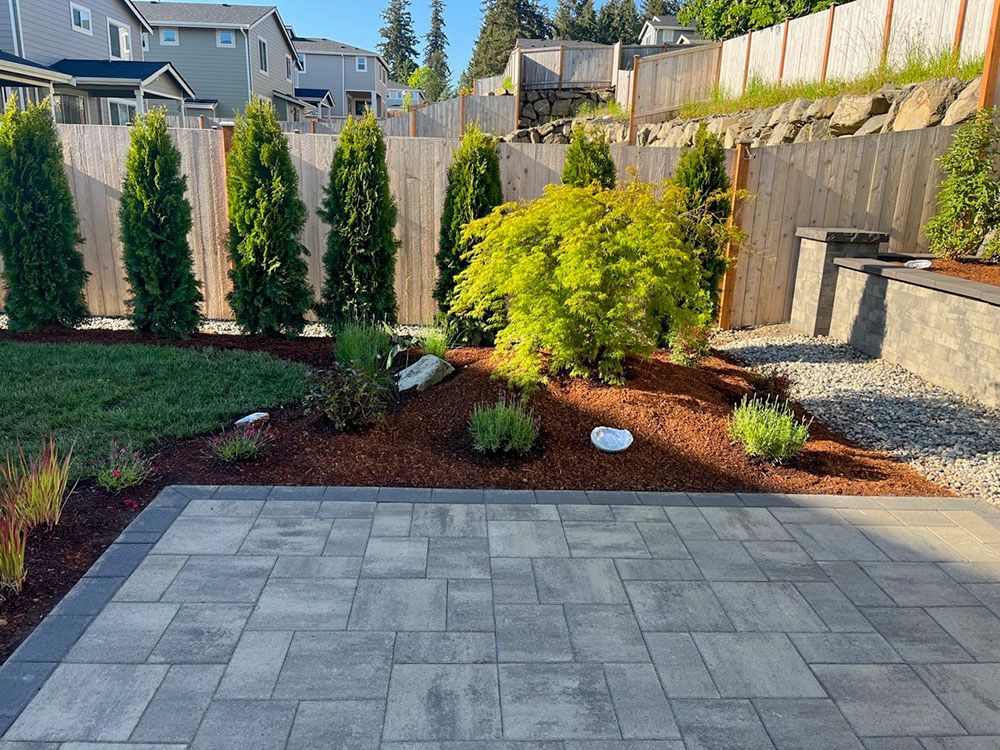 Patio with pavers, mulch, and a landscaped garden with evergreens, a small tree, and a wooden fence.