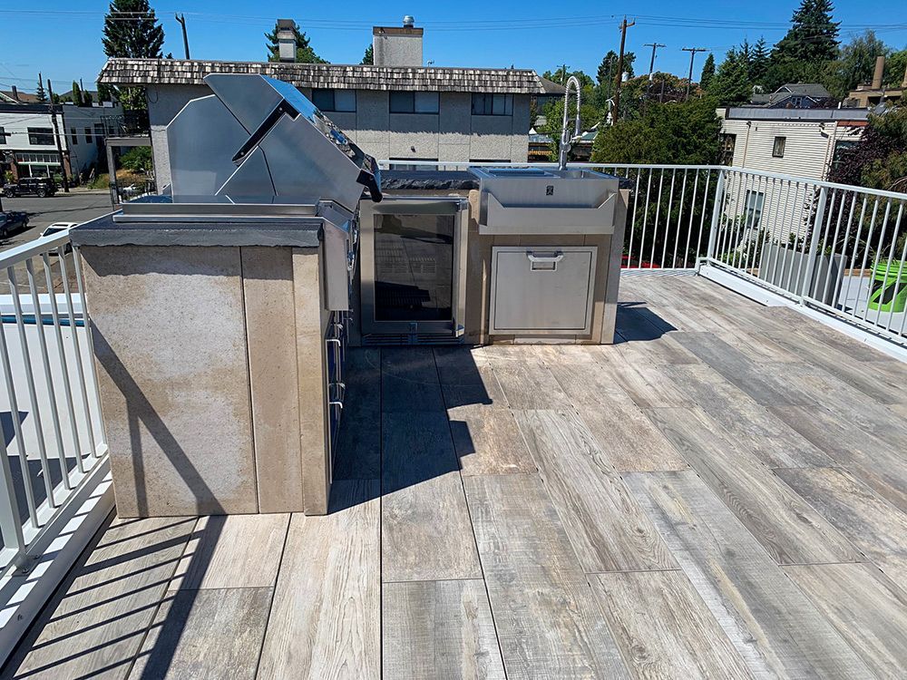 Outdoor kitchen on a rooftop with a grill, storage, and stone countertops. Wooden floor and white railing.