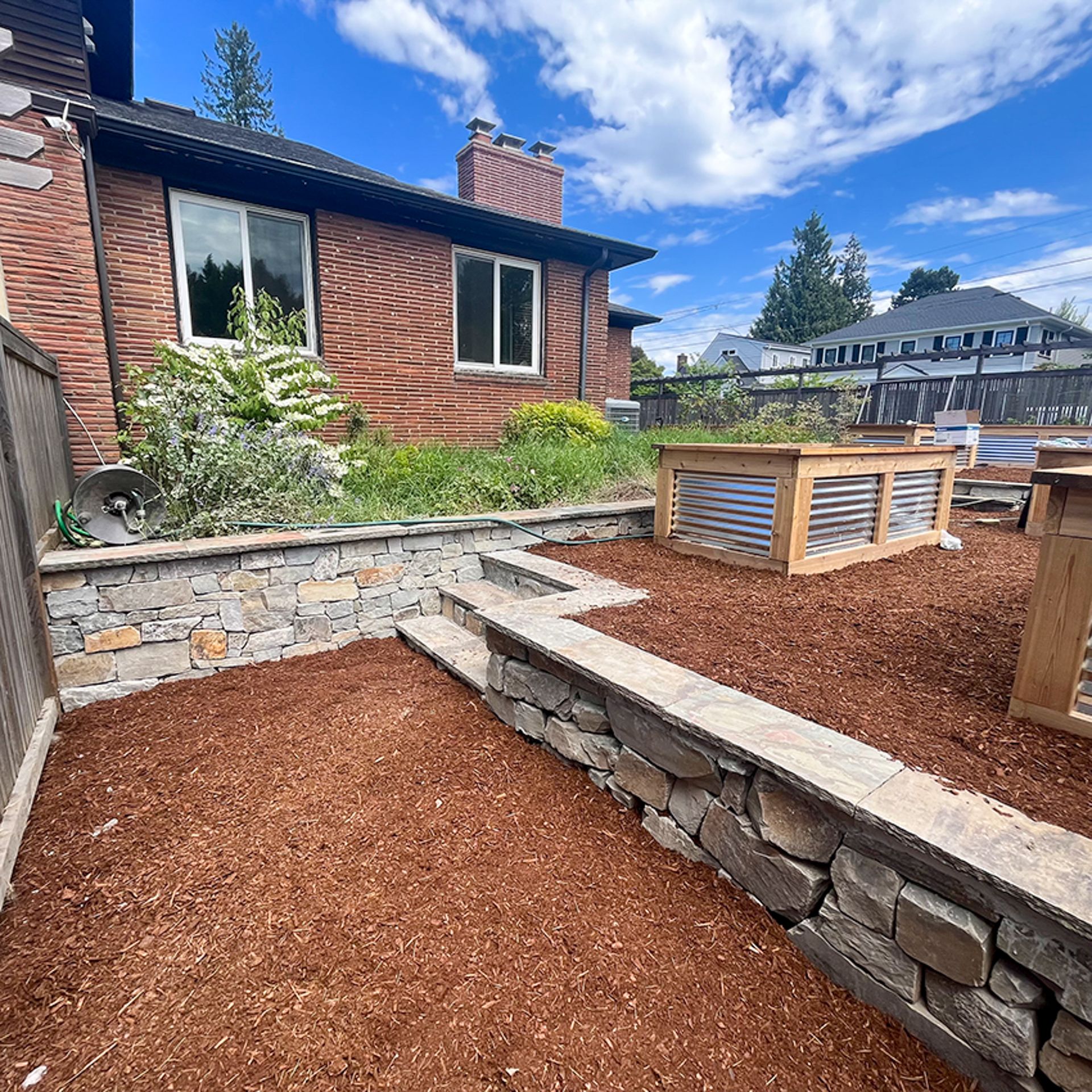 Stone retaining walls with steps leading to a mulched yard, raised garden beds, brick house, blue sky.