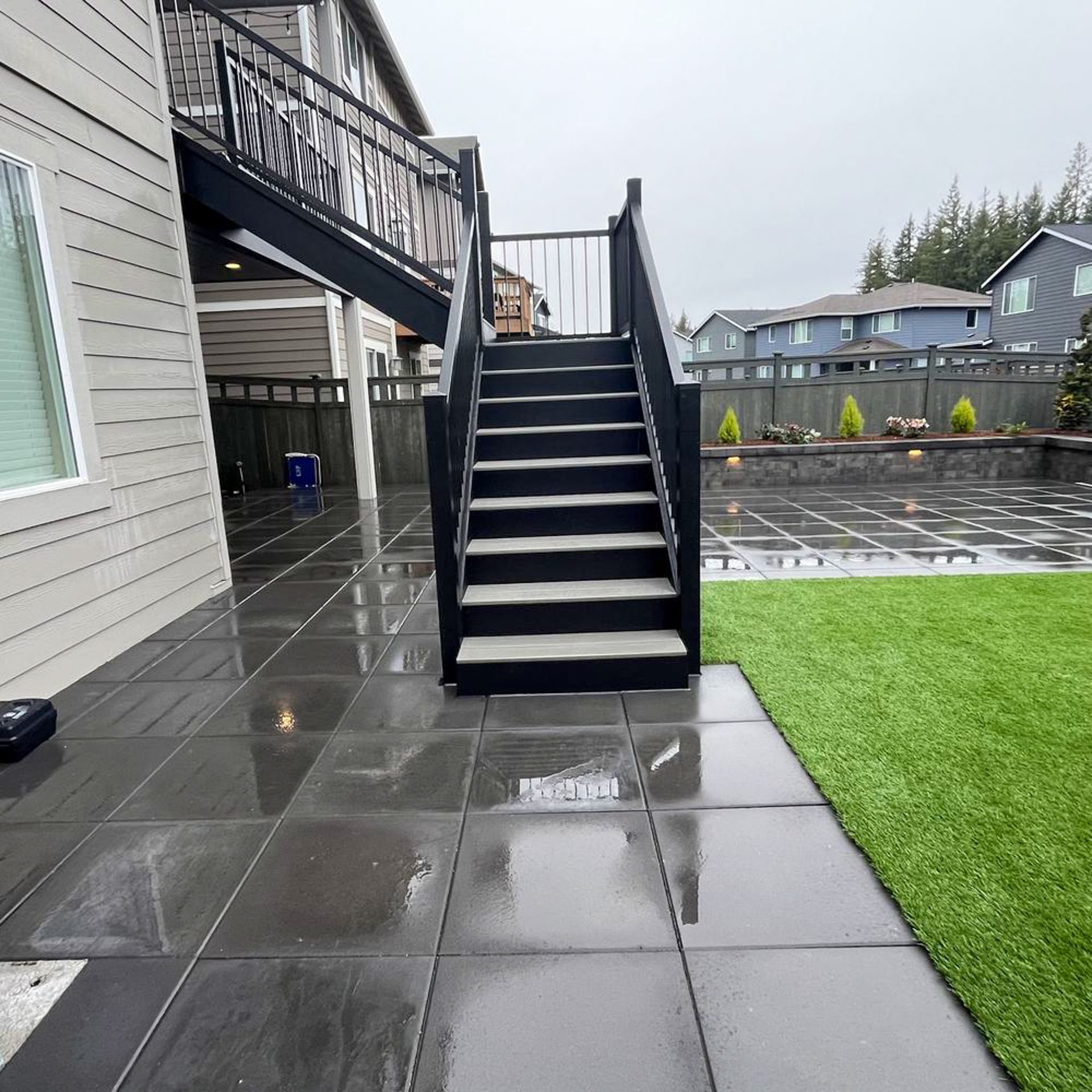 Black outdoor staircase leading up to a deck on a rainy day. Grey patio with green grass.