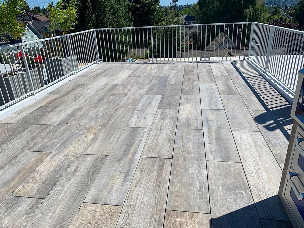 Rooftop deck with light gray wood-look tiles and white metal railing, surrounded by trees and houses.