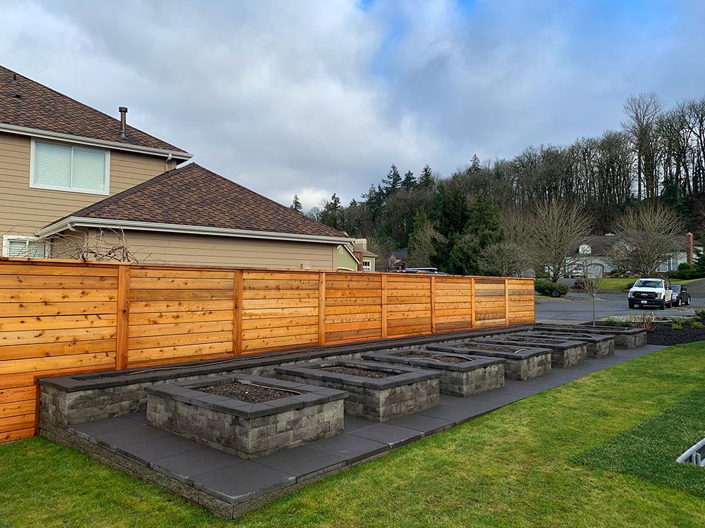 A wooden fence and several square garden beds in a yard. A house and trees are in the background.