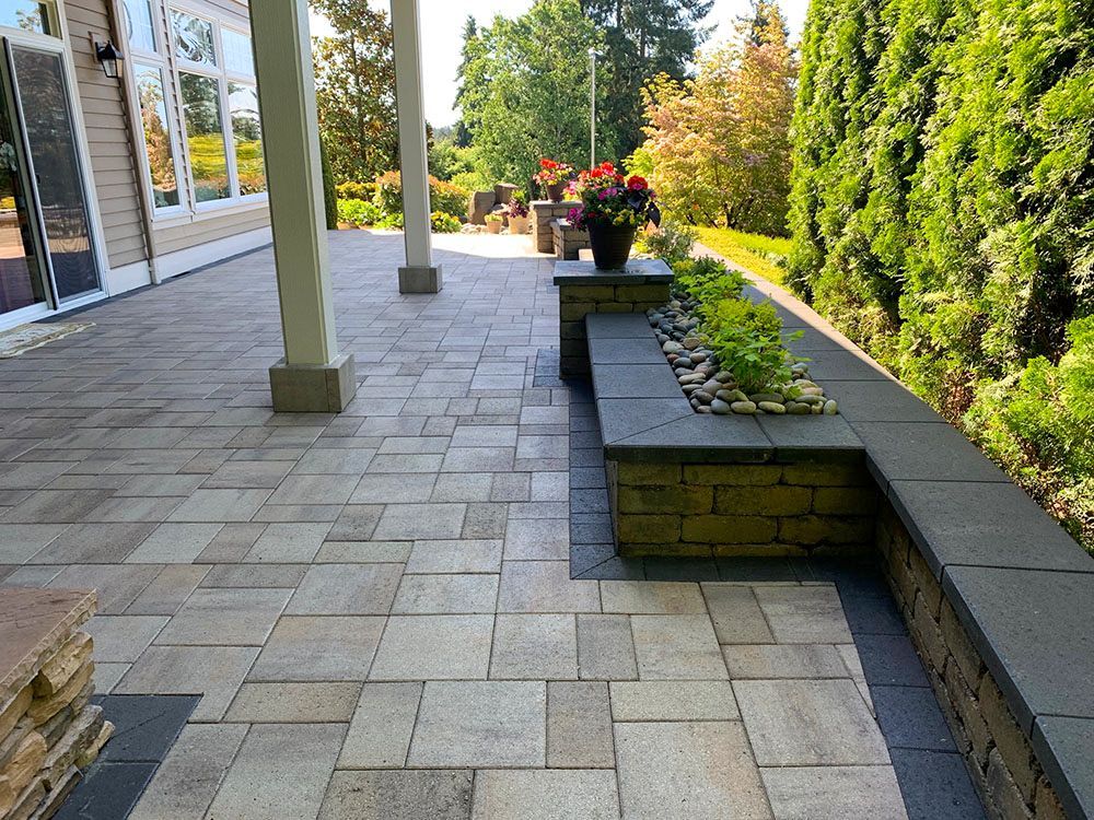 Stone patio with retaining wall, plants, and flowers. Columns support a roof. Sunny outdoor setting.