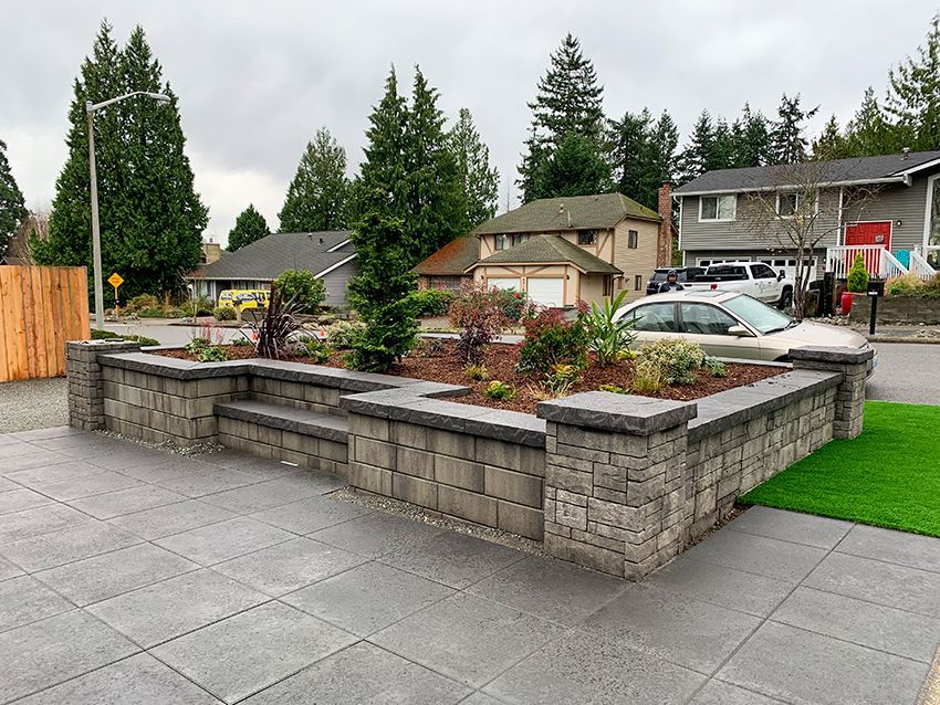 A gray stone retaining wall with a planted garden in front of houses on a residential street.