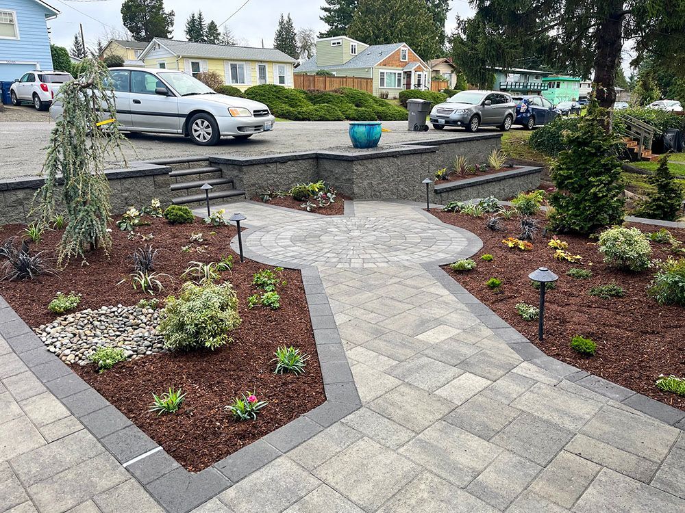 Paver walkway and landscaped yard with raised garden beds, stairs, and parked cars in residential setting.