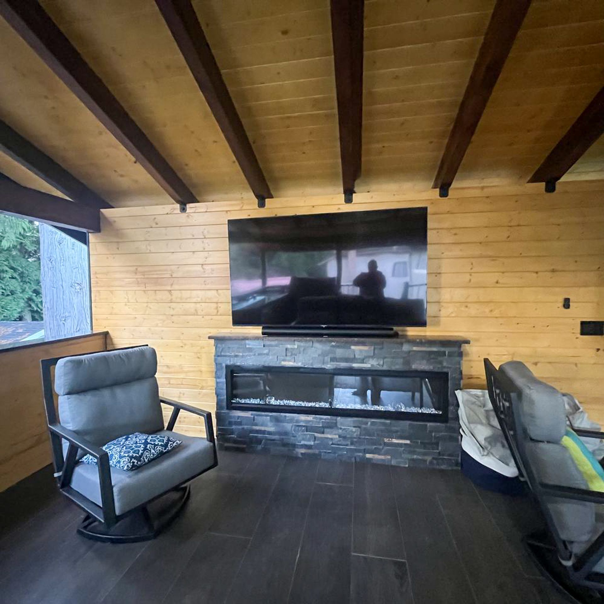 Cozy outdoor room with gray chairs, fireplace, large TV, and wood ceiling.