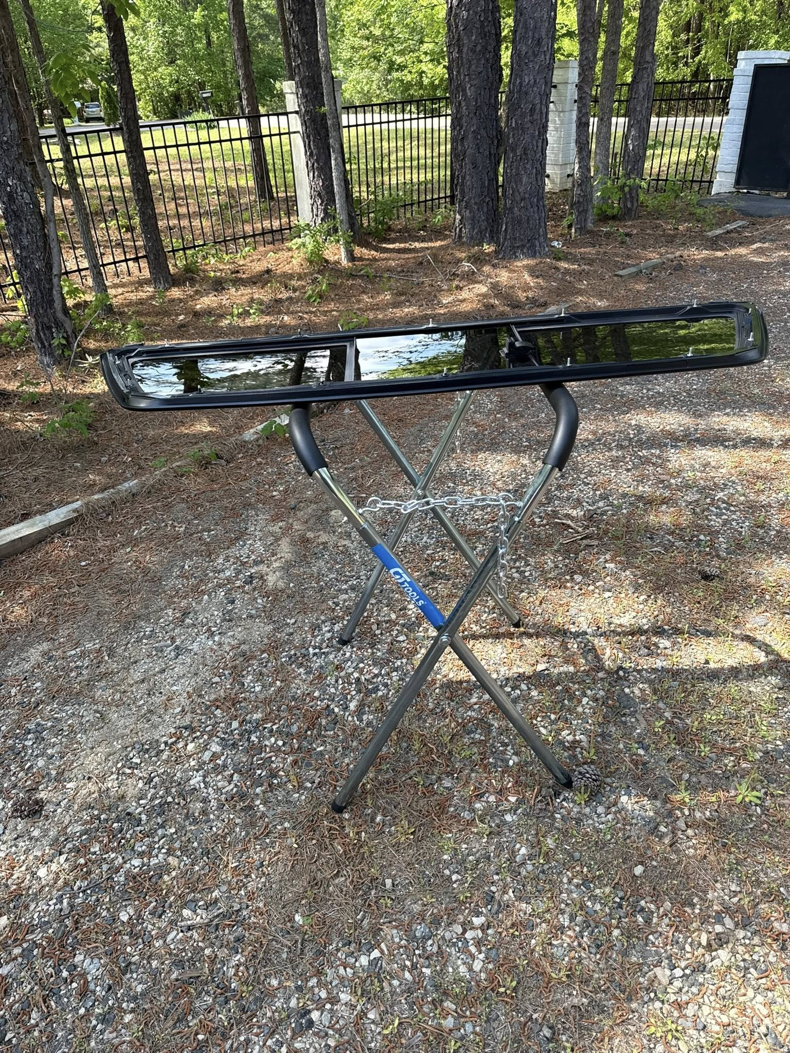 Black keyboard on a folding stand outdoors on gravel, with trees and a fence in the background.