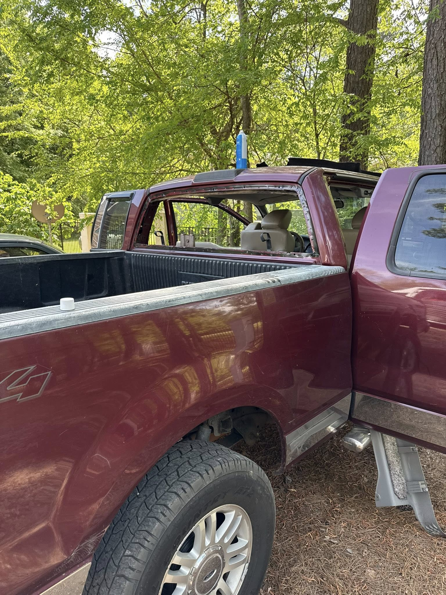 Damaged maroon pickup truck with a crushed cab and broken rear window in a wooded area