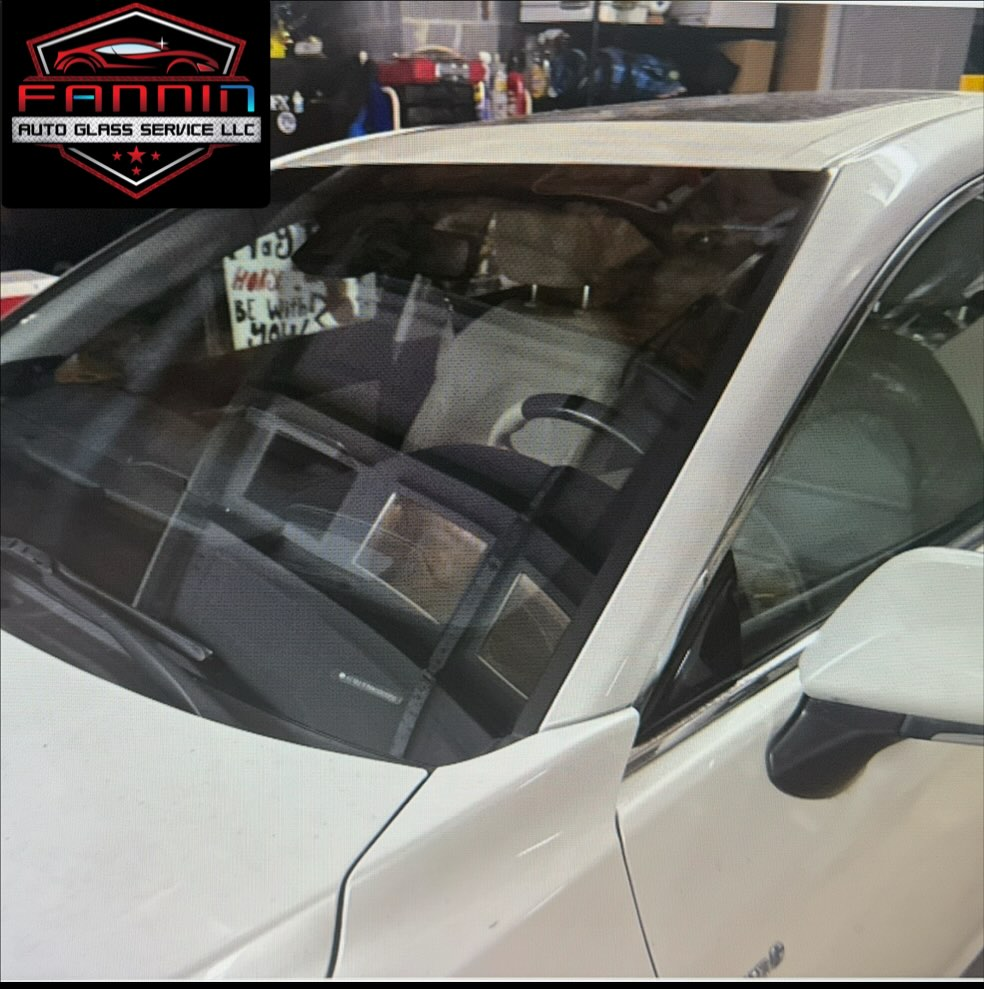 White sedan parked indoors with a windshield sign and auto shop logo reflected on the glass