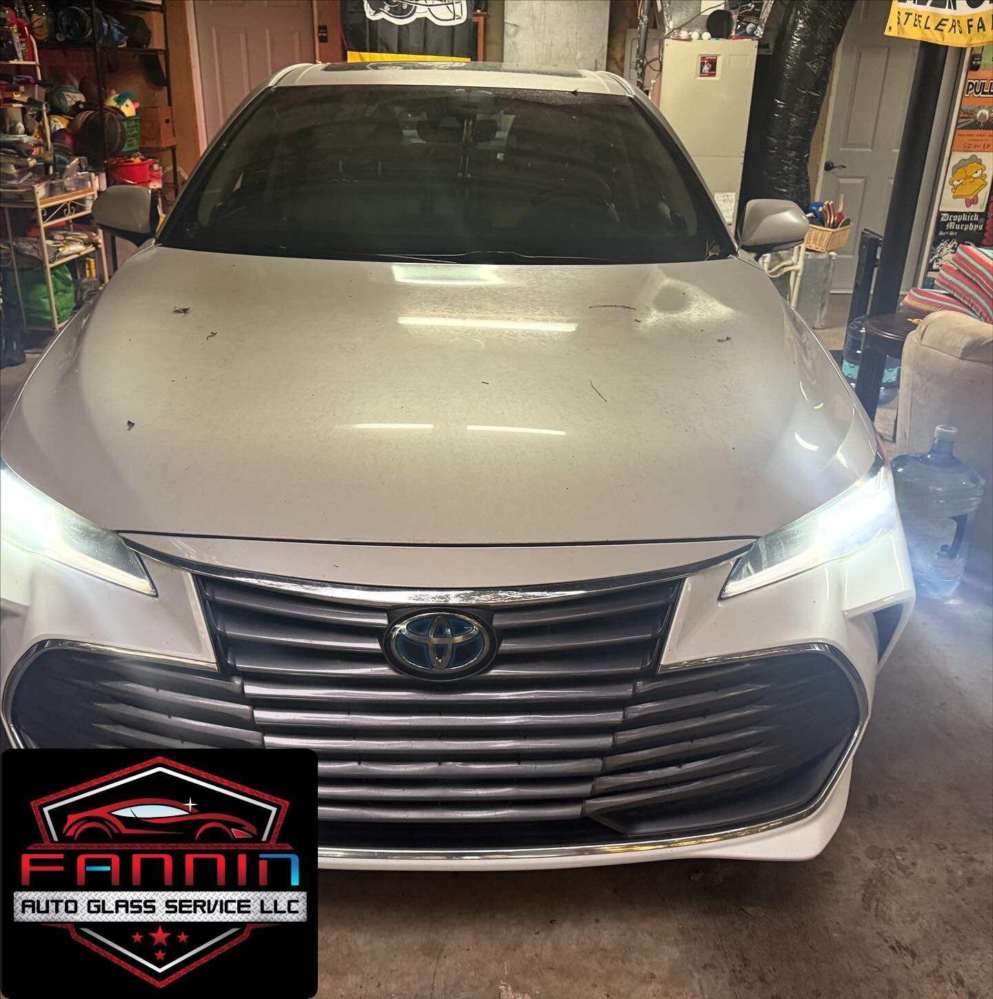 Silver Toyota sedan in a garage, front view with headlights on and a shop logo watermark.