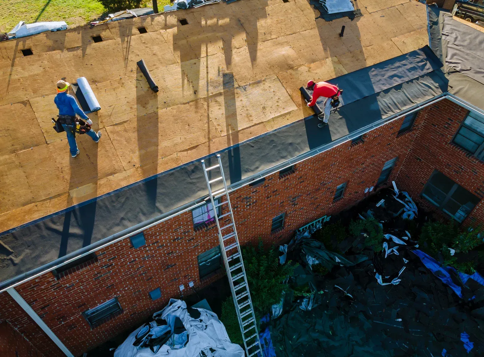 Roofers working on a building roof, installing materials. Ladder and rolled materials are also present.