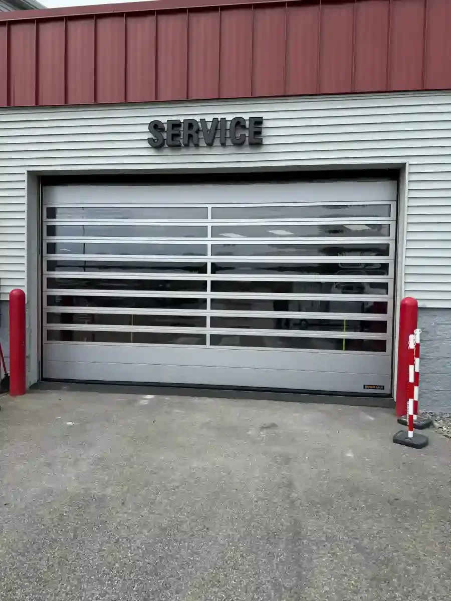 Closed service garage door with horizontal slats, flanked by red bollards, on a gray concrete lot.