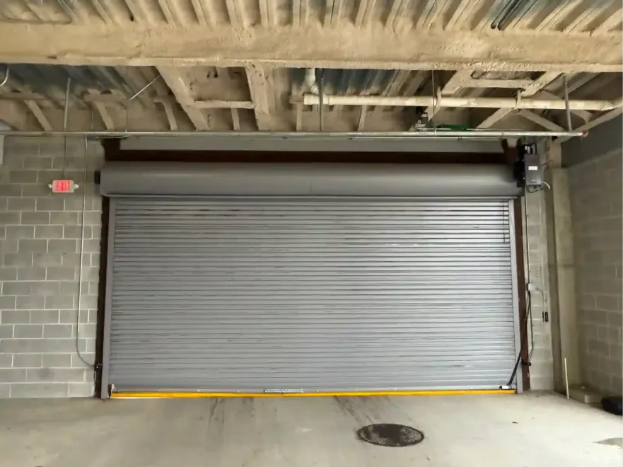 Closed gray roll-up garage door in a concrete parking area with exposed ceiling beams.