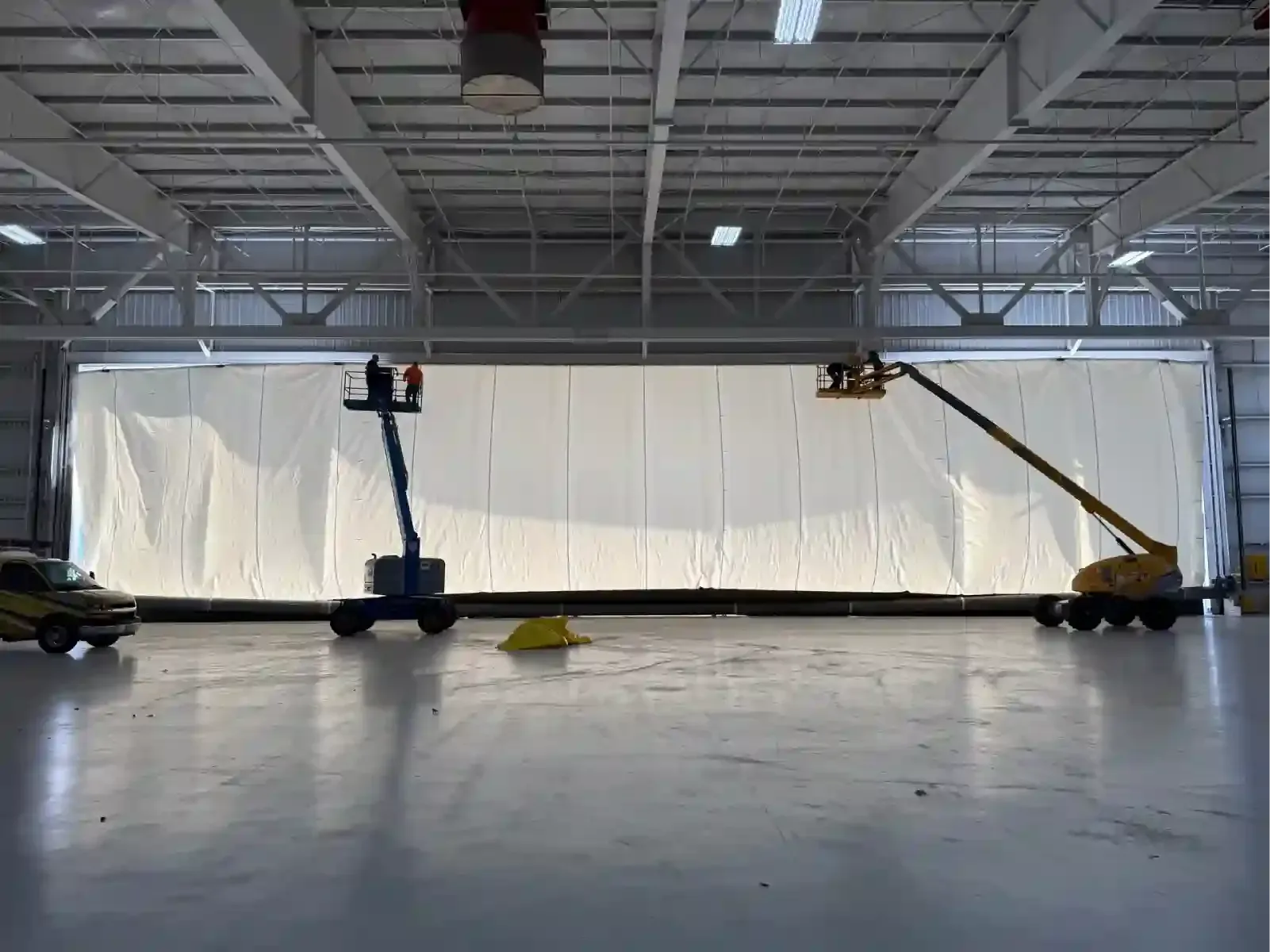 Large industrial hangar with a white barrier, two workers on lifts, and a polished concrete floor