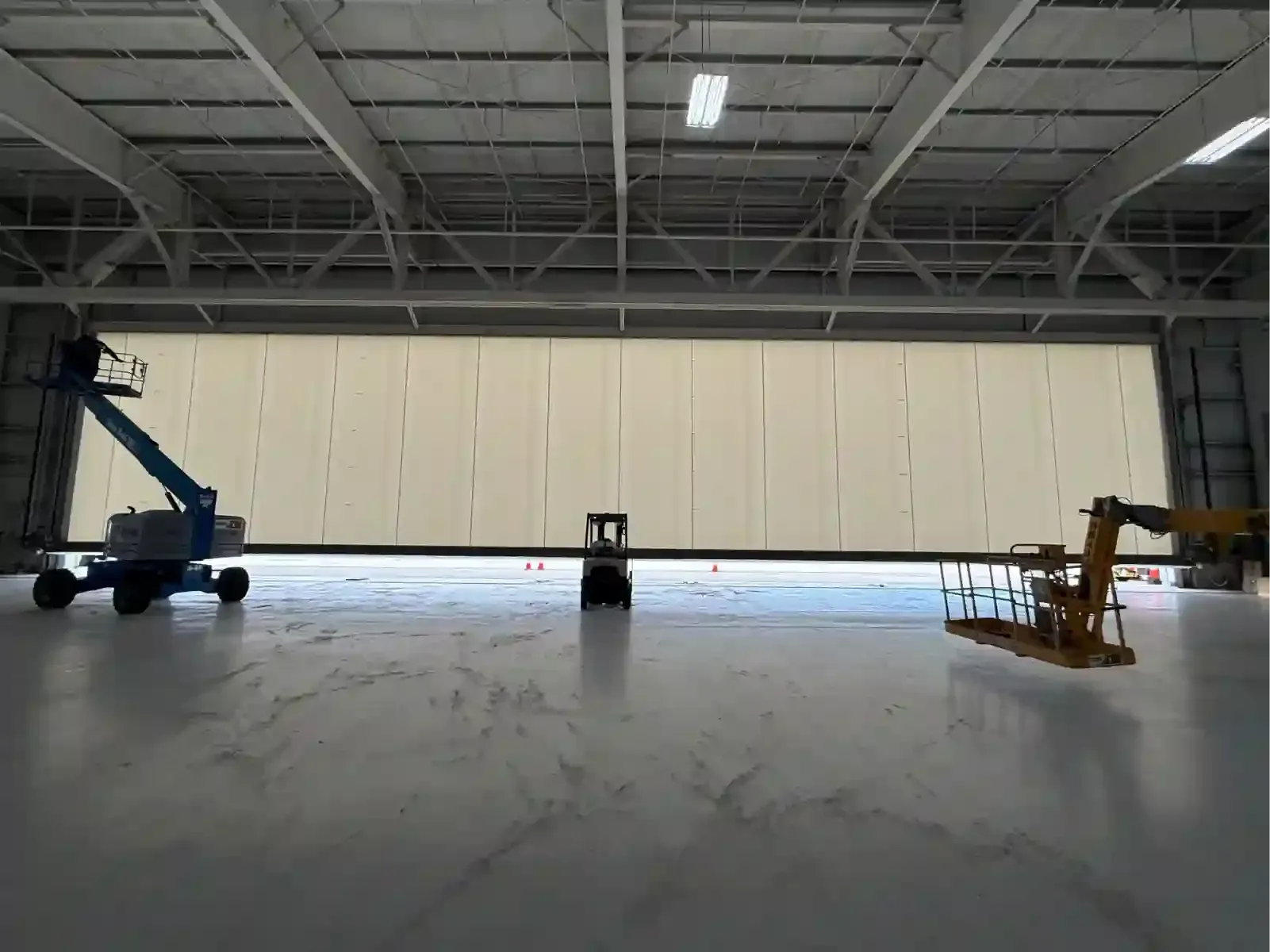 Large industrial warehouse interior with a closed beige hangar door and two boom lifts on the floor.