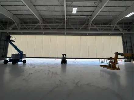 Large industrial hangar with a wide closed door and two lift machines inside, one blue and one yellow.