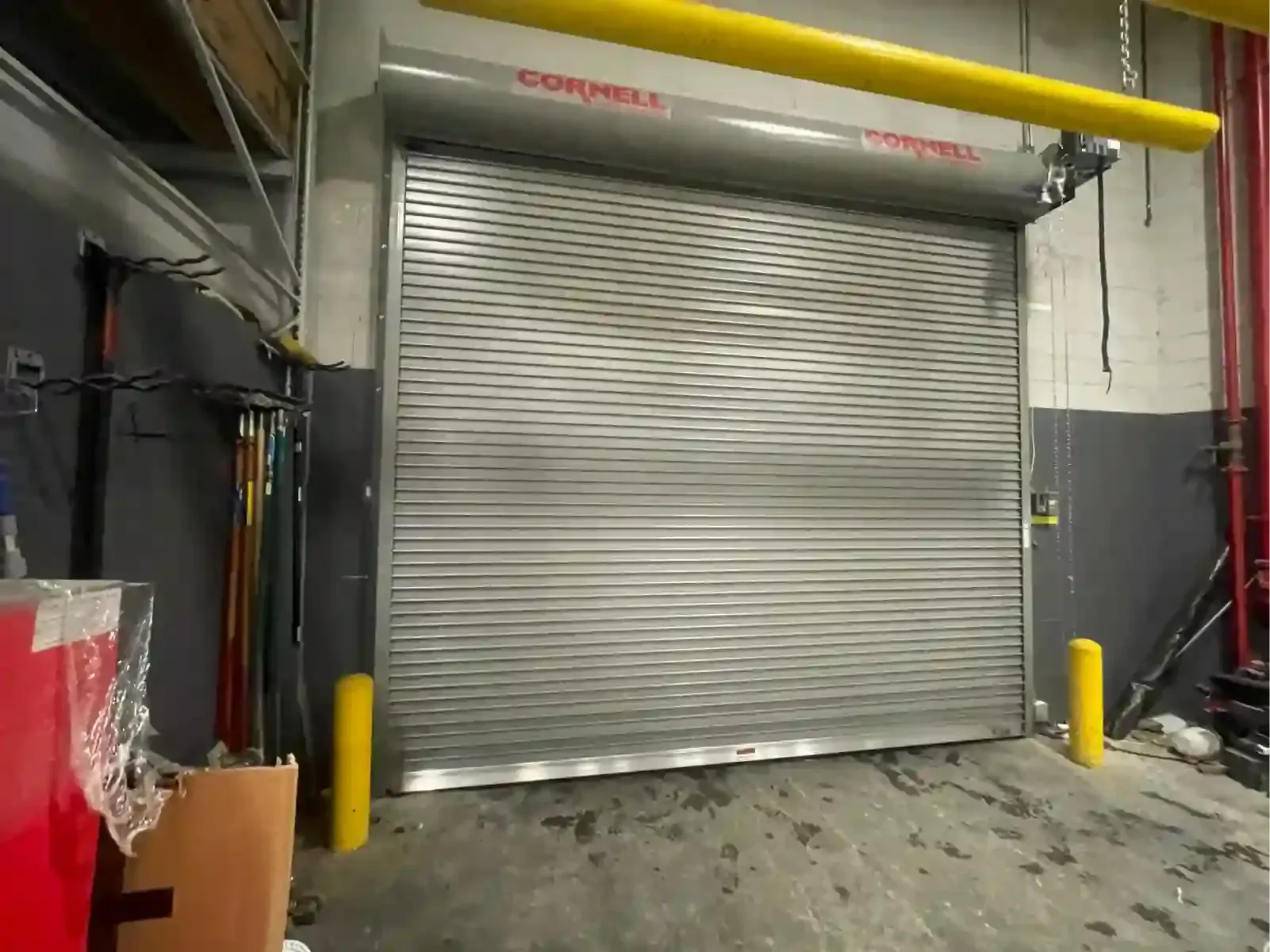 Closed gray roll-up industrial garage door in a warehouse loading area, with yellow protective posts and pipes