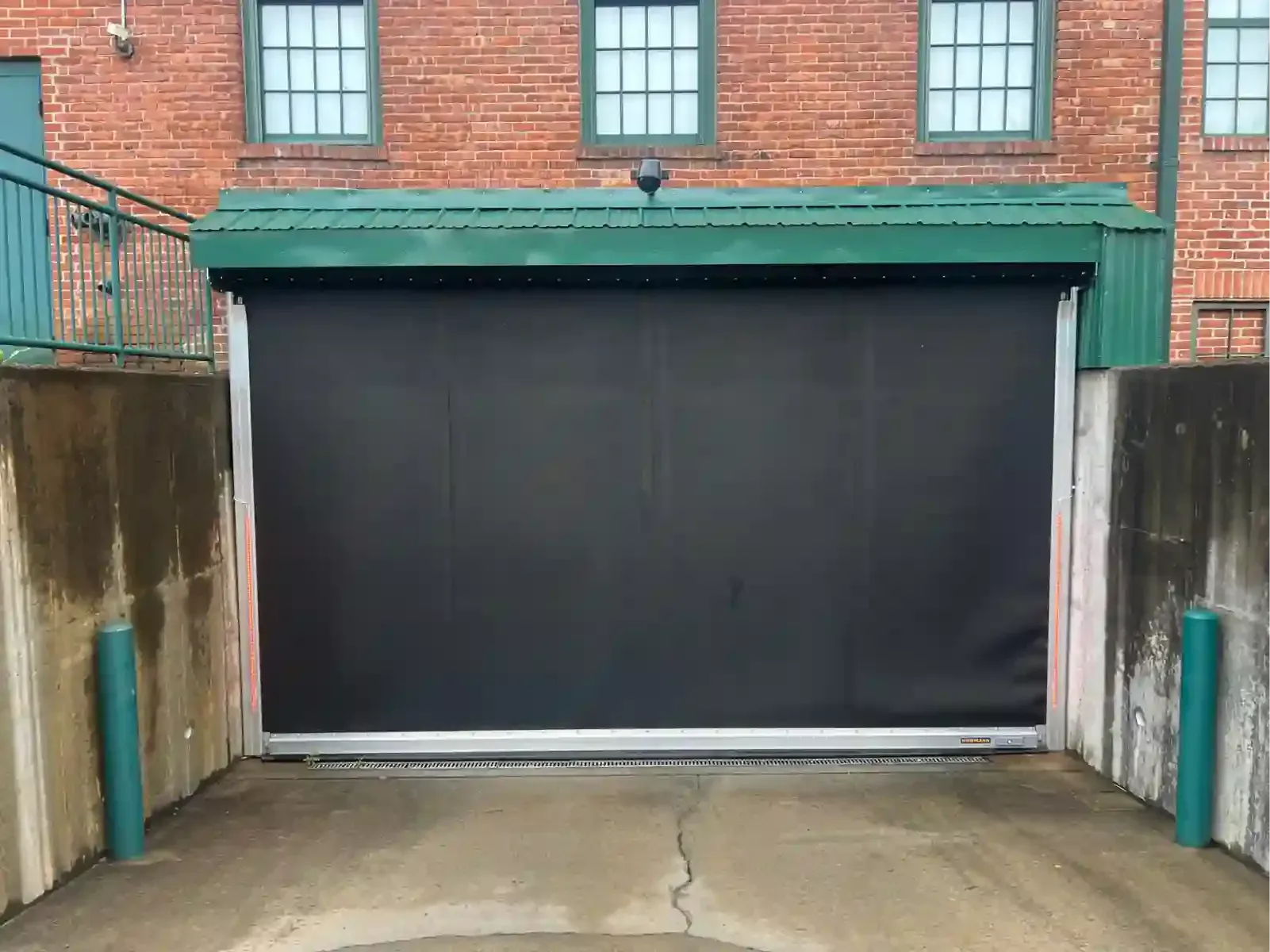 Closed black garage door in a brick building courtyard, framed by green posts and concrete walls