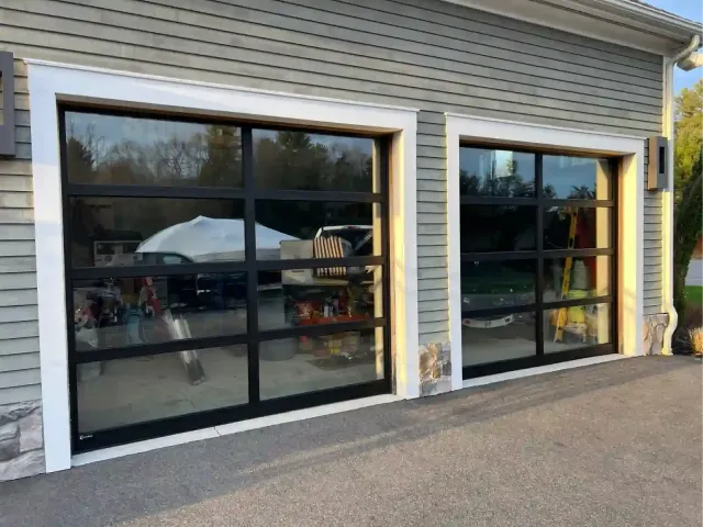 Two black-framed garage doors on a gray-sided building reflect vehicles and a tent outside.