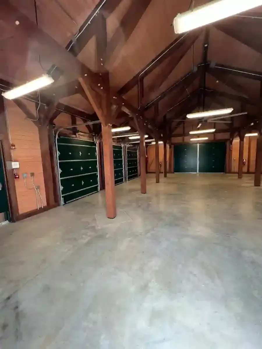 Empty rustic warehouse interior with exposed wooden beams, concrete floor, and green shelving along the left wall