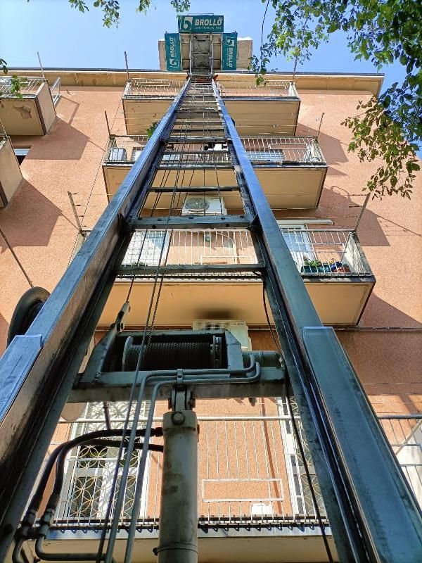 Veduta dal basso di un condominio con vano ascensore esterno e balconi sullo sfondo di un cielo azzurro.