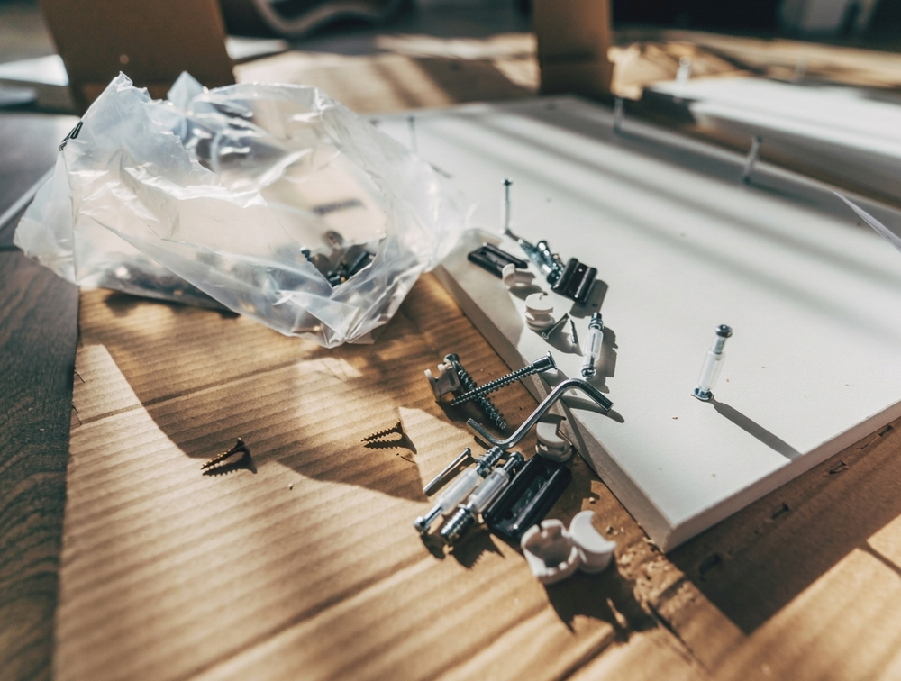 Parti di modellismo e strumenti sparsi su un piano di legno accanto a un pannello bianco e un sacchetto di plastica.